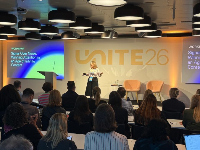 Me on stage at Unite 26, mid-gesture, in a striped top and wide black trousers, speaking to a full room. The screen beside me reads "Workshop: Signal Over Noise: Winning Attention in an Age of Infinite Content".