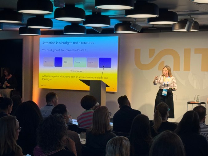 A slide reading "Attention is a budget, not a resource. You can't grow it. You can only allocate it." Coloured blocks for HR, IT, CEO, Comms and Payroll fill one working day. I'm on stage beside it, explaining.