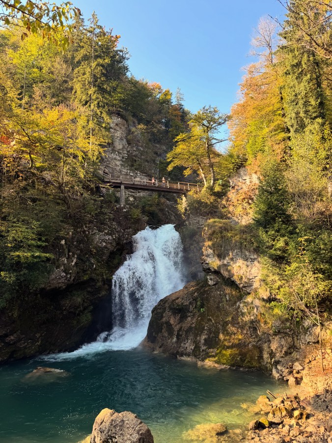 A white waterfall pours between mossy rocks into a deep blue-green pool. A wooden bridge crosses high above it, framed by golden autumn trees and blue sky.