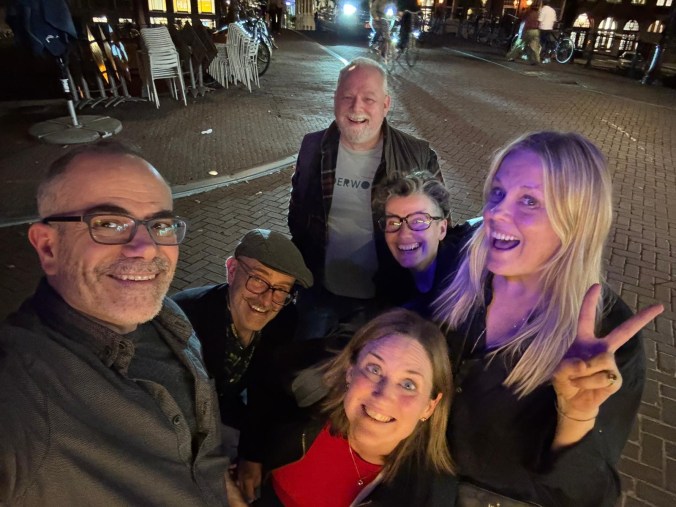 A late-night street selfie of six friends crouched together on cobbles, all grinning widely. One woman flashes a peace sign, and bikes and stacked café chairs sit in the background.