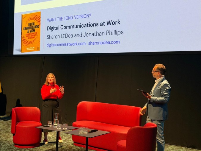 Sharon, in a red top, gestures mid-answer on a stage with red sofas as a moderator holding a tablet listens. Behind them, a large screen shows the cover of Digital Communications at Work and the words "Want the long version?"