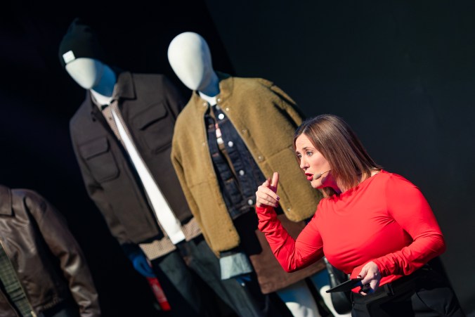 Sharon on stage in a red top and headset mic, leaning forward and pointing emphatically, with faceless white mannequins in autumn jackets standing behind her.