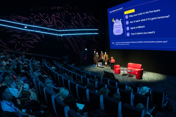 A packed cinema-style auditorium watches Sharon on a spotlit stage. The giant screen reads "Test it before you need it", with a four-step to-do list and the quote "A crisis is not the moment to discover whether communication systems work".