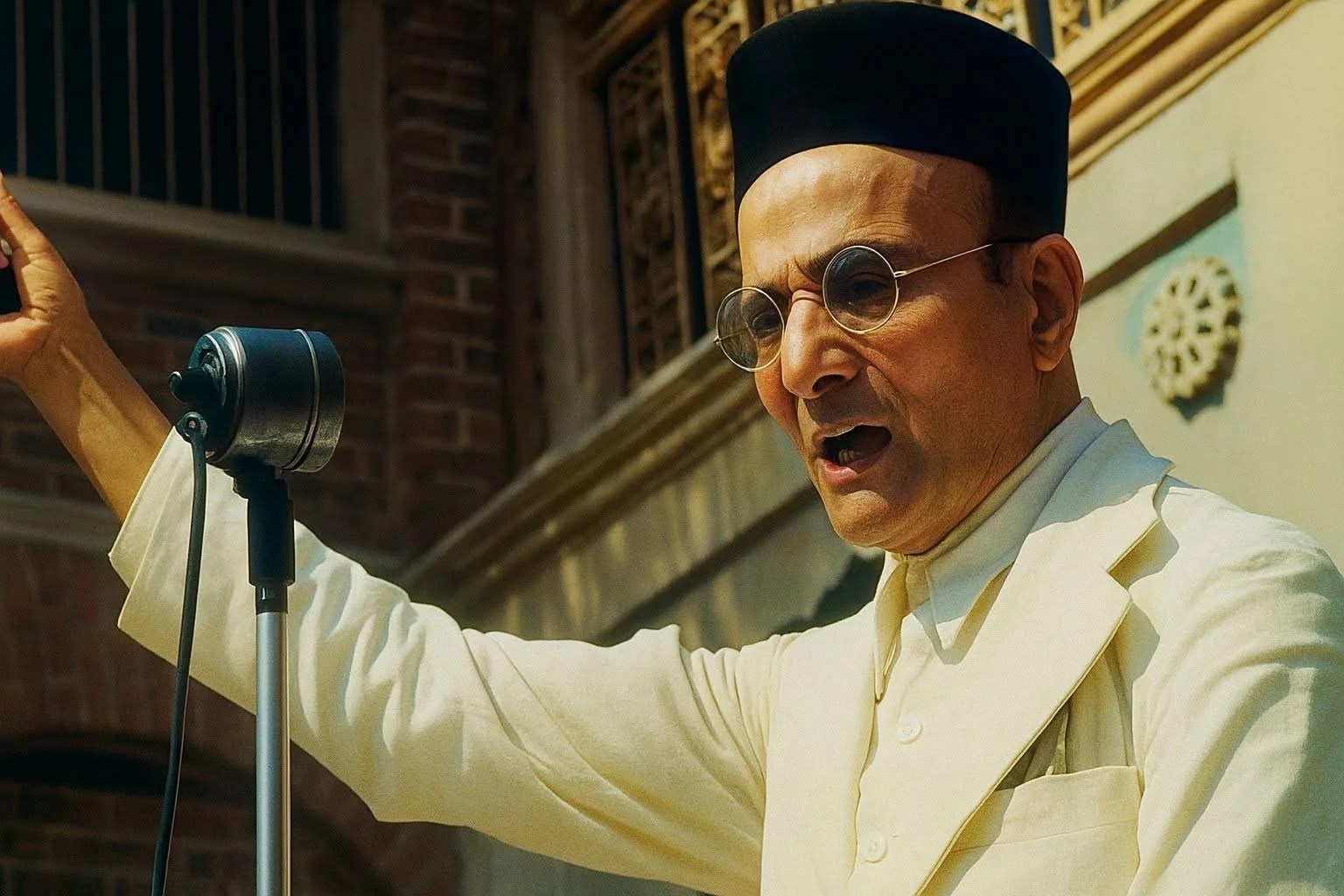 A historical figure in traditional Indian attire and round glasses, wearing a black cap, passionately addressing a gathering through a vintage microphone.