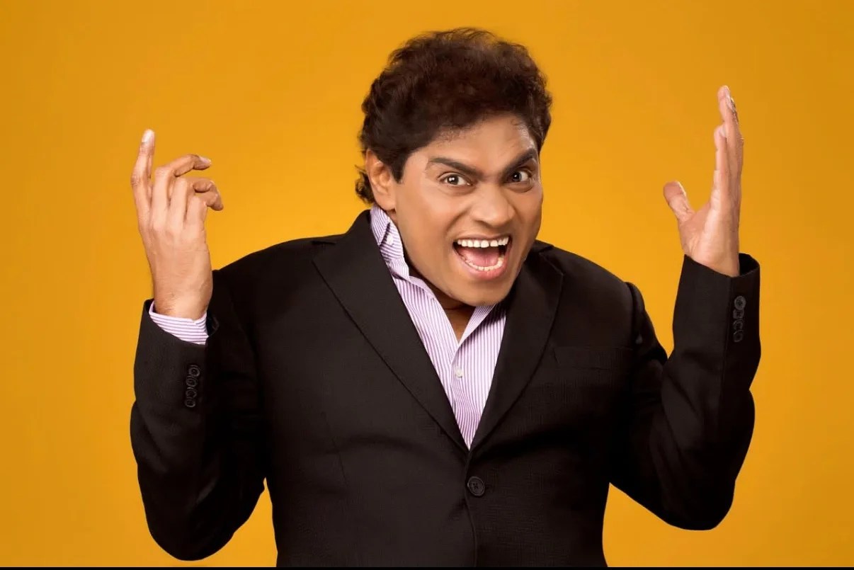 काले सूट में हँसते और मज़ाकिया अंदाज़ में हाथ उठाए हुए मशहूर कॉमेडियन जॉनी लीवर का चित्र।