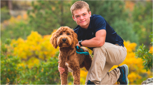 A teenage boy kneeling by a golden doodle dog outdoors.