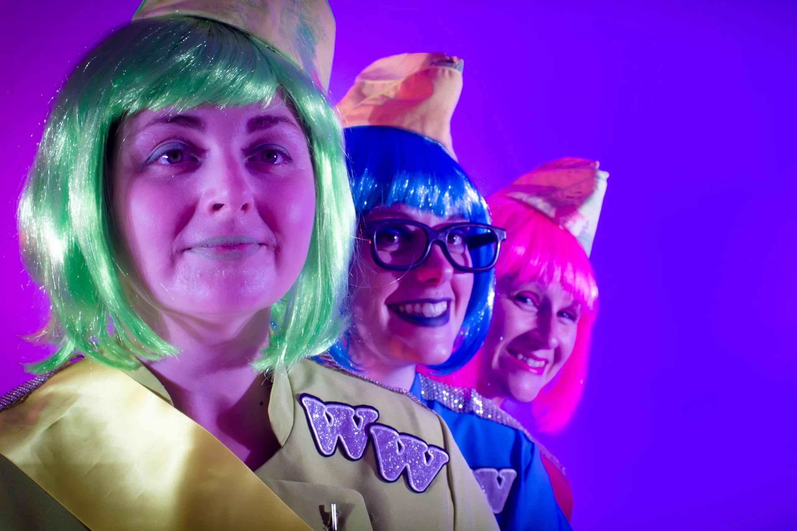 Captain Wonderweb, Engineer Spectech and Lieutenant Fishingnet. Three people in costume stand in a diagonal line, each wearing a brightly coloured bob-cut wig (Wonderweb - green, Spectech - blue, and Fishingnet - pink) and matching outfits. They smile under vivid purple lighting, with the closest figure looking directly at the camera.