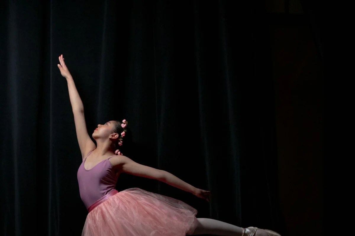 A ballerina in a pink tutu reaching upward in an arabesque on a dark stage