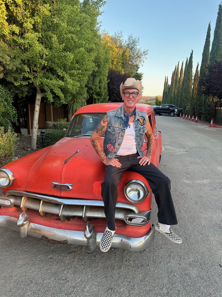 Man with tattoos in a denim vest and cowboy hat sits on the hood of a rusty red vintage car, smiling at the camera outdoors.