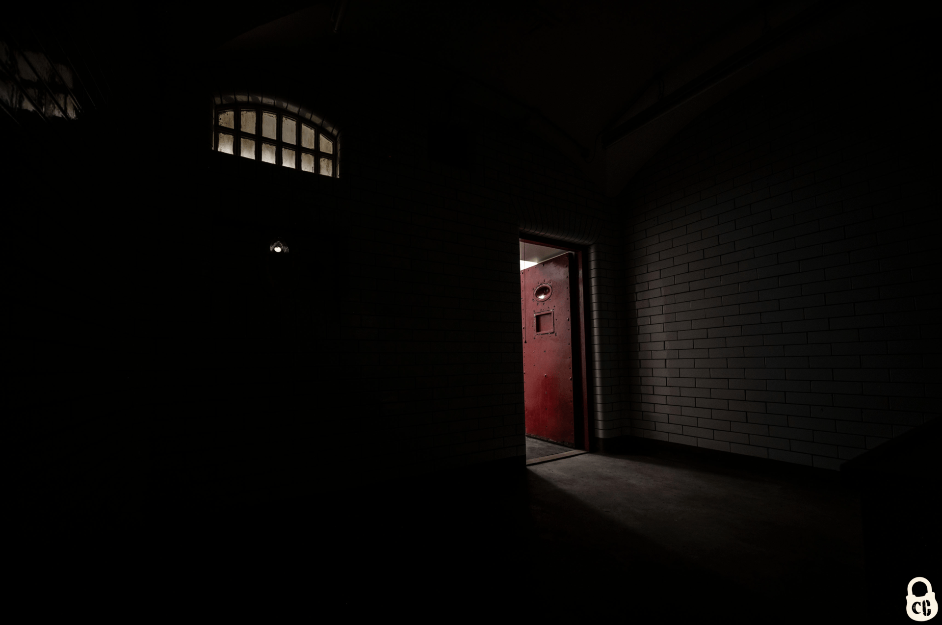 A dark Victorian cell with light coming in through a small window and an open red door.