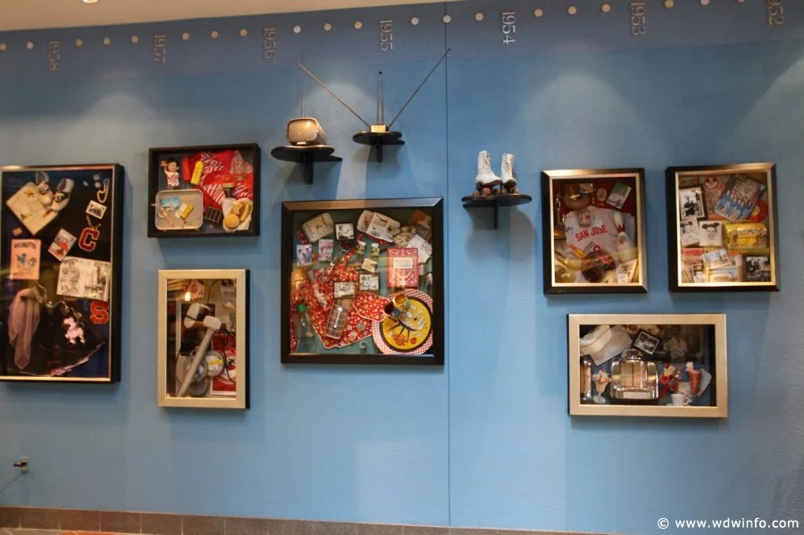 Blue wall gallery with multiple framed collage displays of photos, postcards, and memorabilia arranged in a grid variety of sizes and frames.