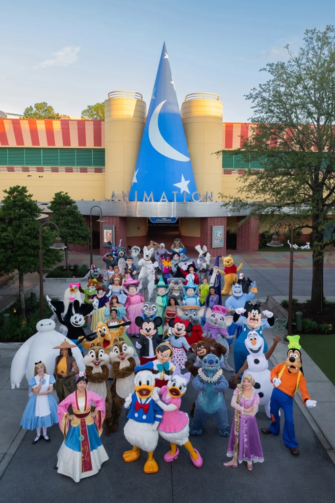 Large crowd of Disney character mascots posing together outside an Animation building at a theme park.