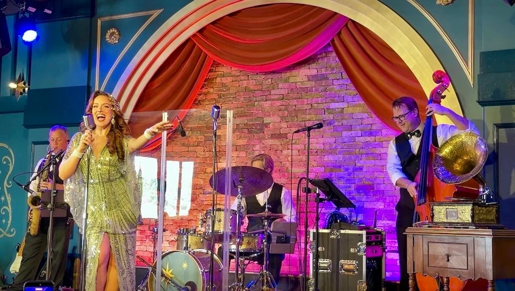 Female singer in a glittering silver gown performs center stage with a live band on a brick backdrop.