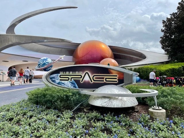 image0 - SiDisney Mission Space sign with a ringed sculpture and colorful planets outside a futuristic ride building; visitors walk nearby.
