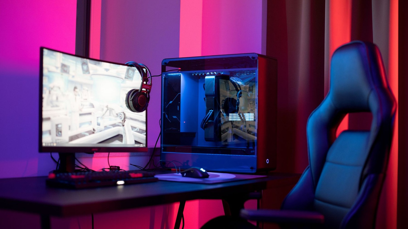 Ambiente gamer iluminado por luzes coloridas, com computador, monitor panorâmico e cadeira.