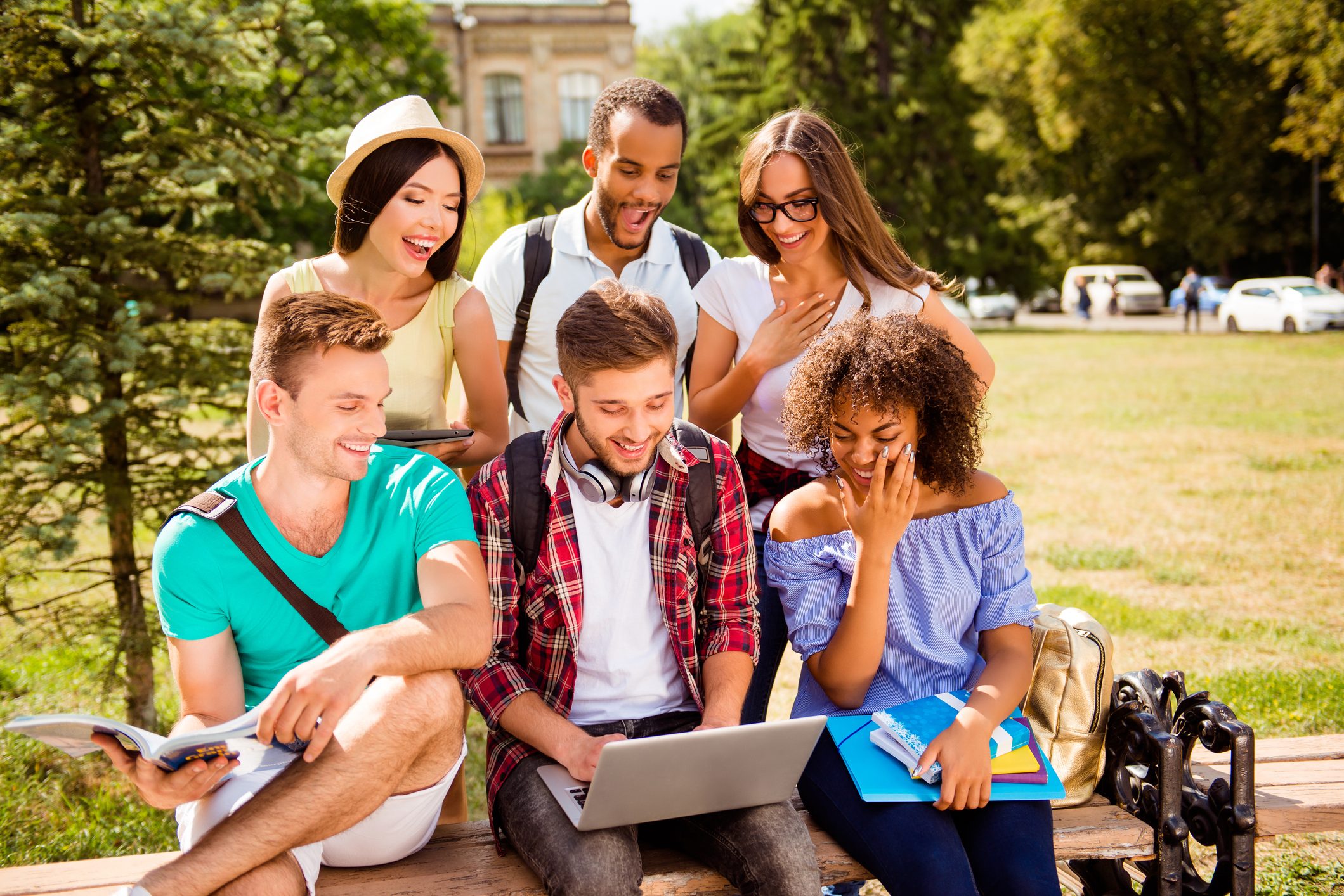 A group of six young adults, diverse in appearance, gathered outdoors on a university campus. They are sitting on a bench and standing around, smiling and laughing while looking at a laptop screen. Some are holding books and notebooks, dressed casually, with backpacks nearby, suggesting a study session or a shared moment of entertainment. The background features green trees, grass, and a historic-looking building.