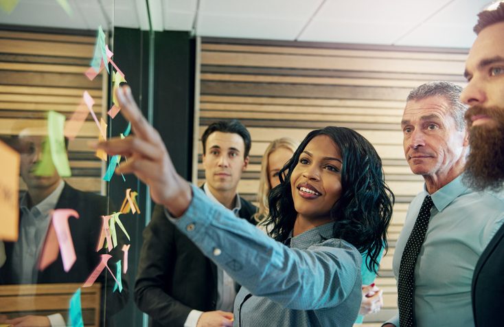 Business team collaborating in a meeting, with a woman leading a brainstorming session using sticky notes on a glass board.