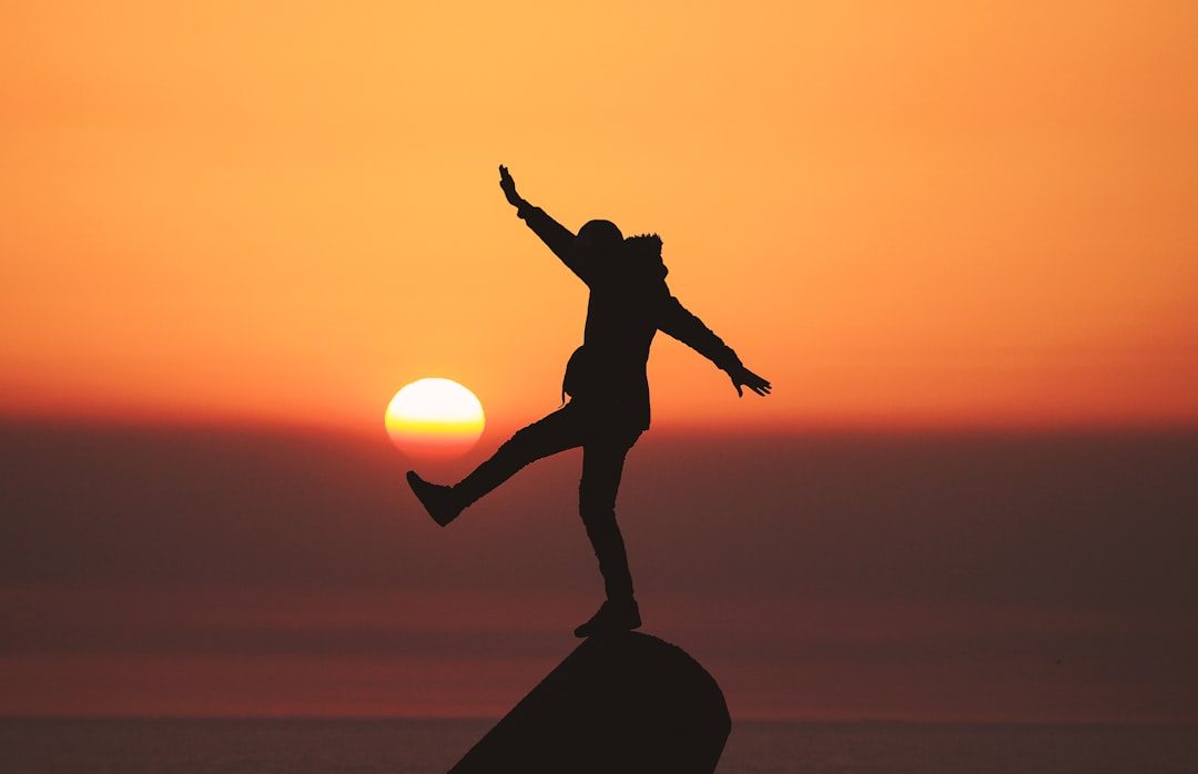 A silhouette of a person balancing on one foot atop a rock, with arms outstretched and one leg lifted, against a sunset sky. The sun appears directly behind them, casting a warm orange glow across the horizon.