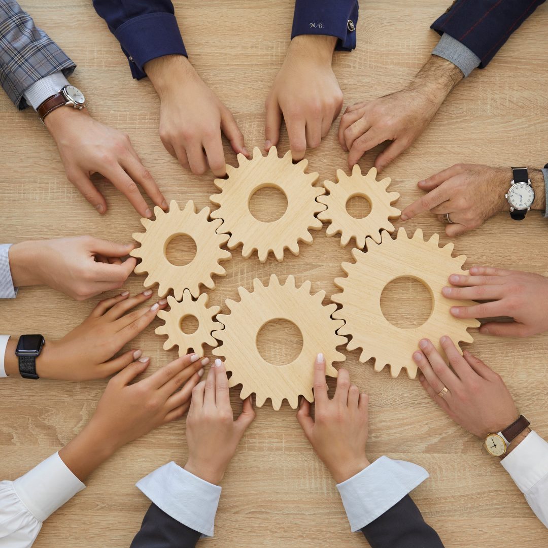 A top-down view of multiple hands working together to connect wooden gears on a table, symbolizing teamwork, collaboration, and synergy in a professional setting.