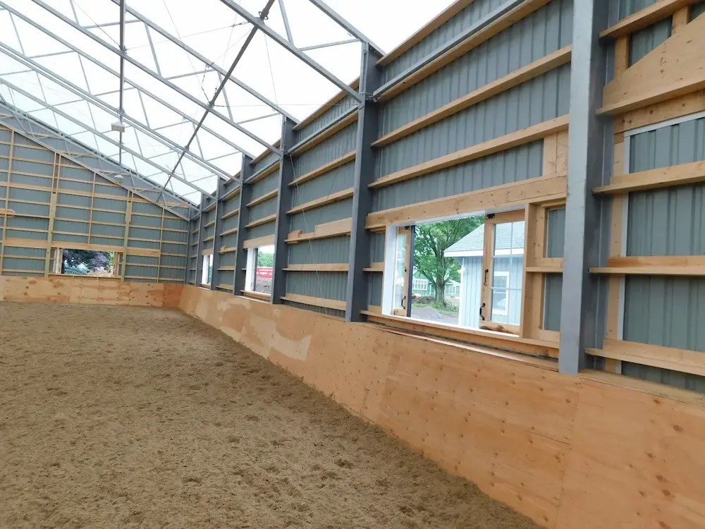 Commercial covered horse arena interior showcasing clear-span steel frame engineering and large custom viewing windows by an equestrian construction firm
