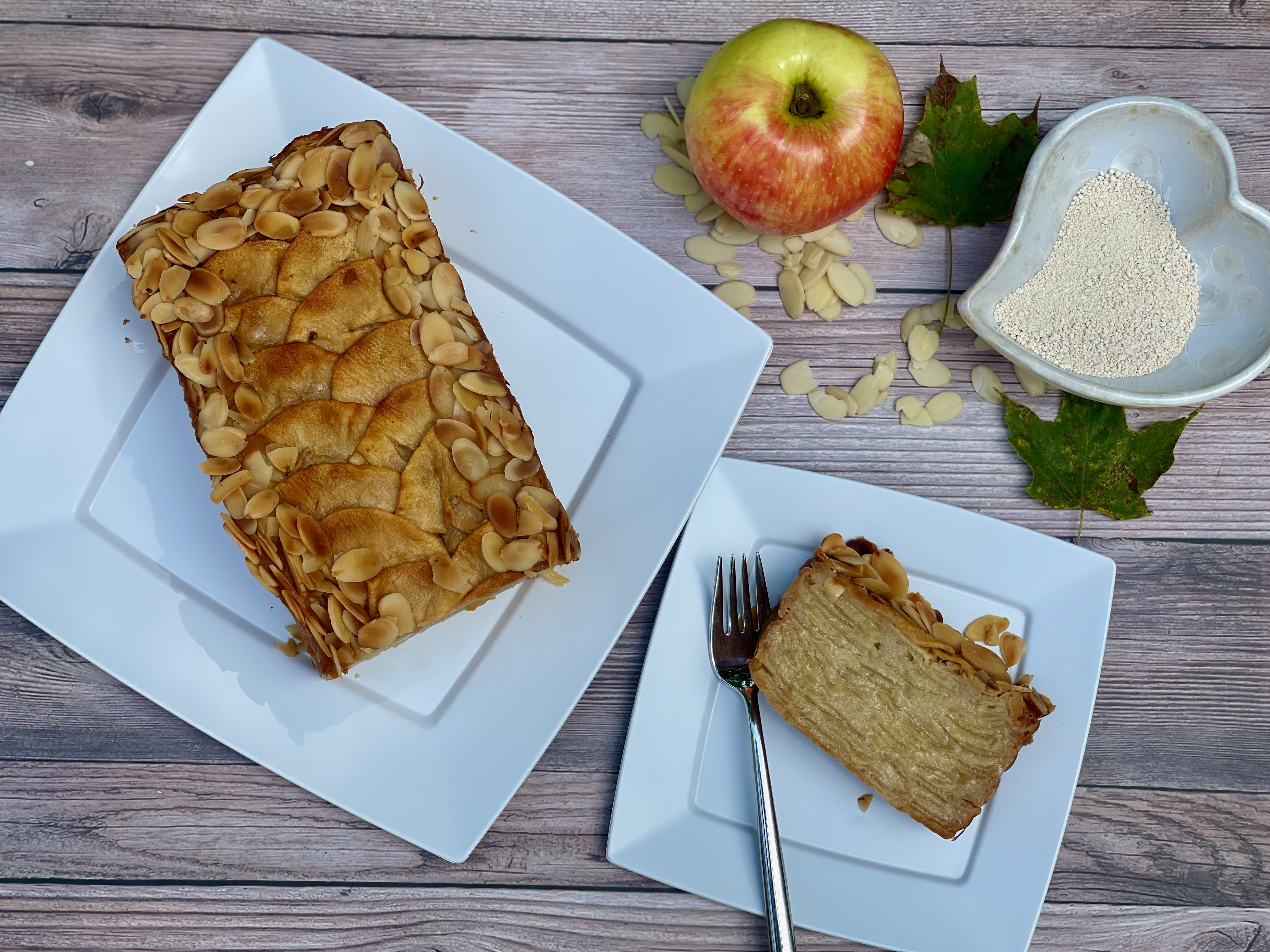 A slice of gluten-free invisible apple maple cake, maple sugar, an apple, almonds and a plate with the cake