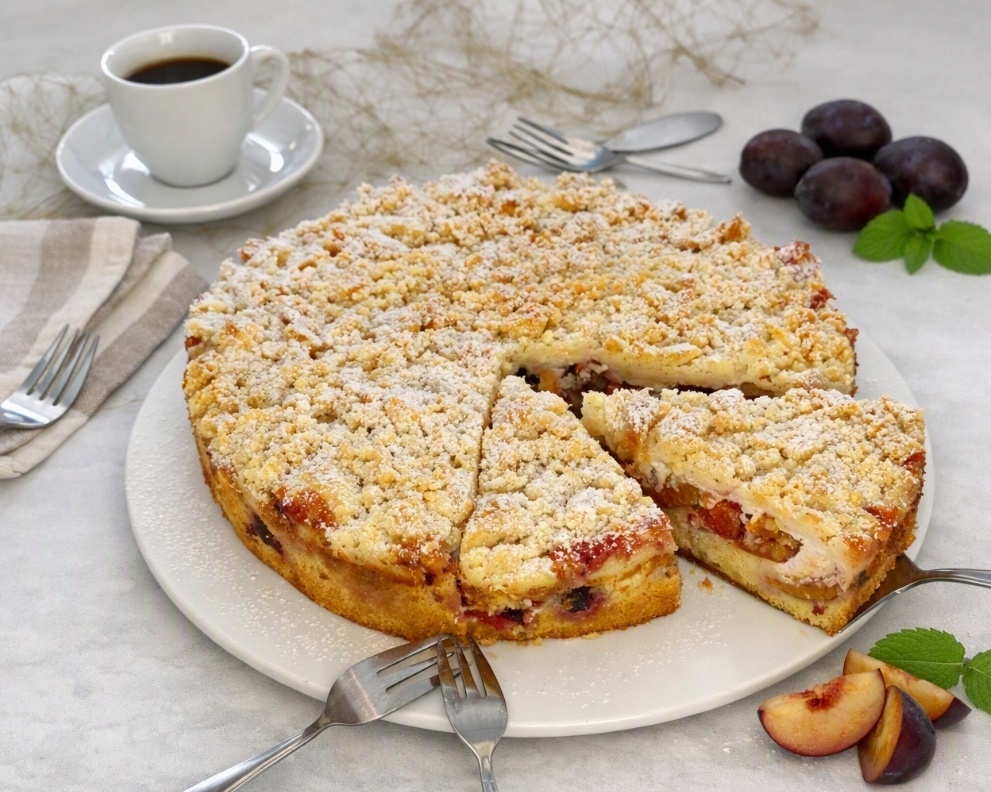 Saftiger Pflaumenkuchen mit Streusel gebacken in einer Springform, angeschnitten, im Hintergrund eine Tasse Kaffee und Pflaumen als Dekoration