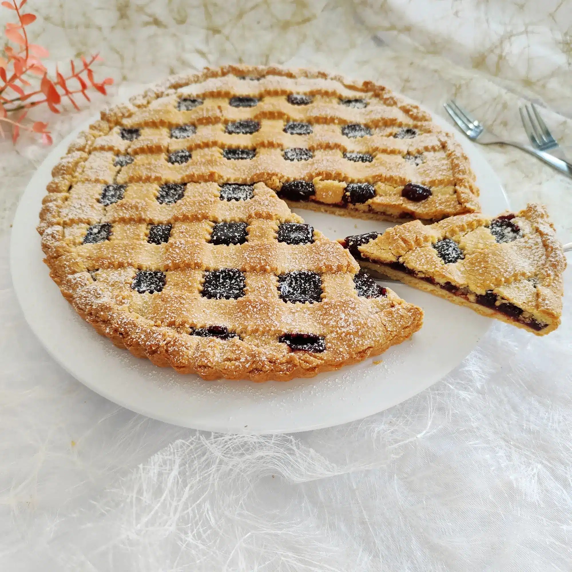 ruchtige Tarte mit selbstgemachter Marmelade, Gitter aus Mürbeteig und Puderzucker, angeschnitten im Vordergrund und zwei Kuchengabeln im Hintergrund