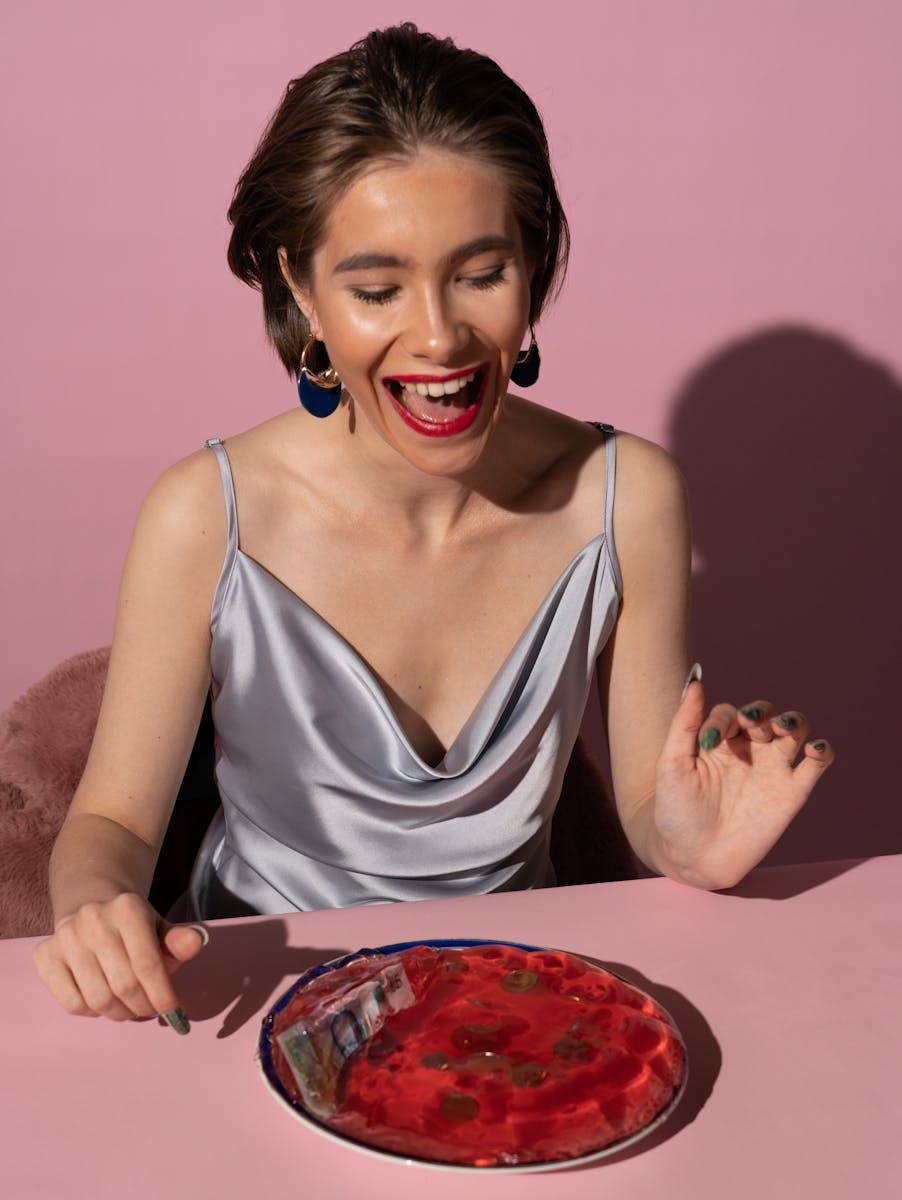 A young woman in a silk dress excitedly views a dessert with a playful expression, set against a pink background.