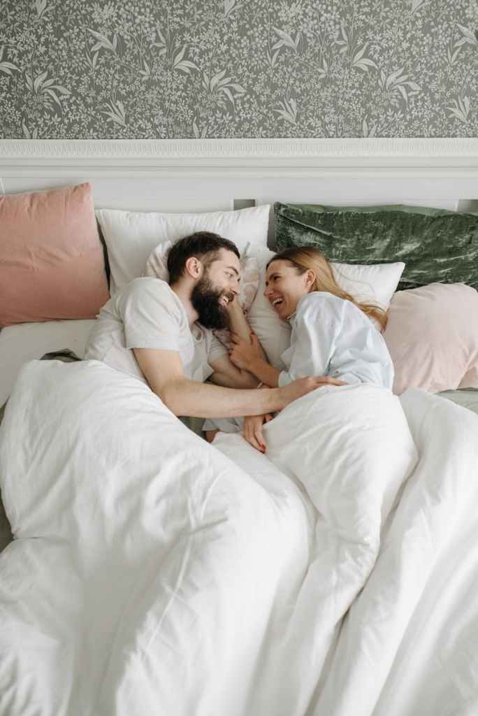 Happy couple smiling and bonding in a cozy bed, enjoying a relaxed morning in a stylish bedroom setting.