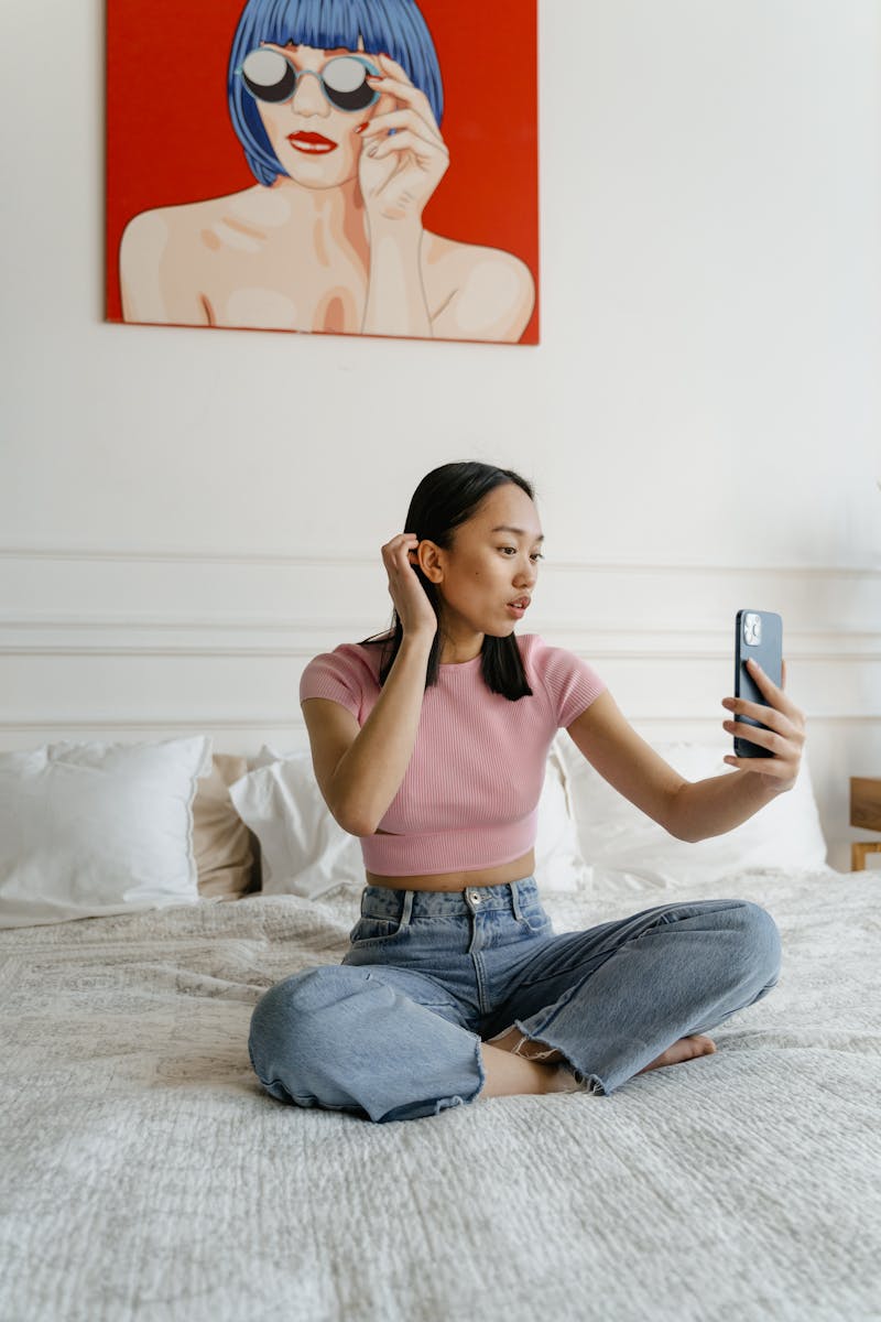 A young woman sitting on a bed taking a selfie in a contemporary styled bedroom.