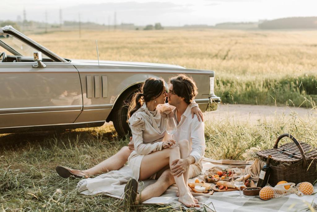 A romantic couple shares a picnic in a scenic field beside a classic car, capturing love and tranquility.