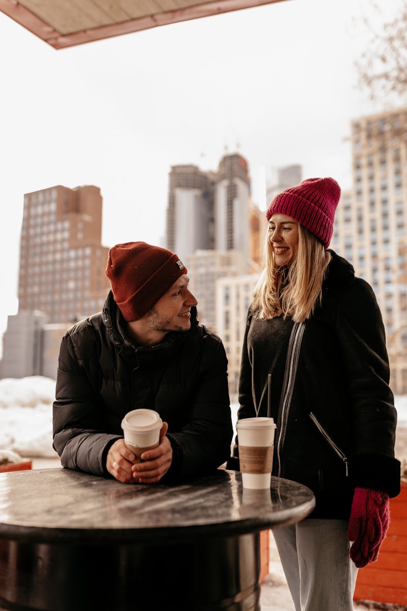 Smiling couple in warm clothing enjoying coffee outdoors in a winter urban setting.
