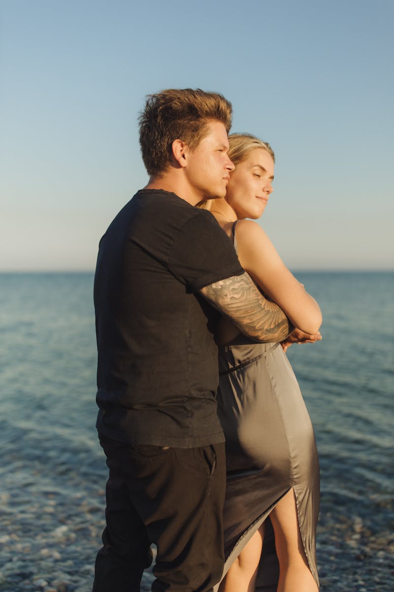 A couple embraces on a pebbled beach with the ocean and sunset in the background, capturing a romantic moment.