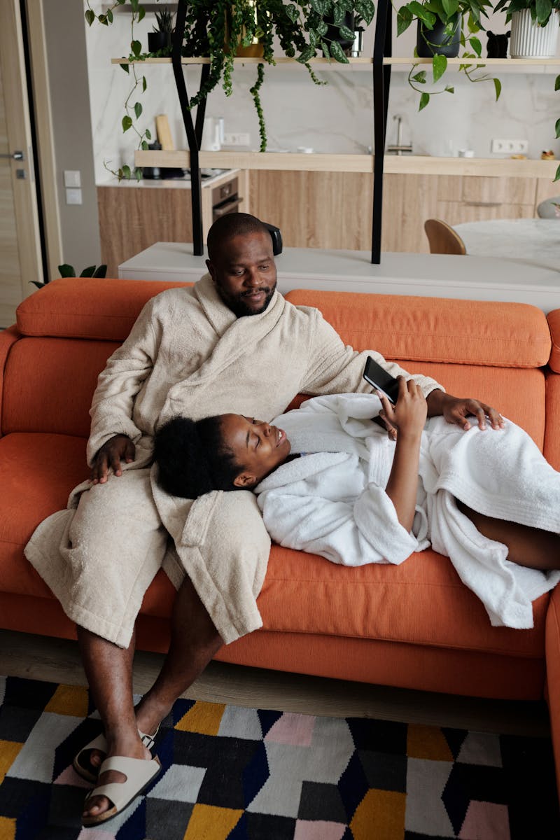Smiling couple in bathrobes using smartphone while relaxing on a cozy sofa indoors.