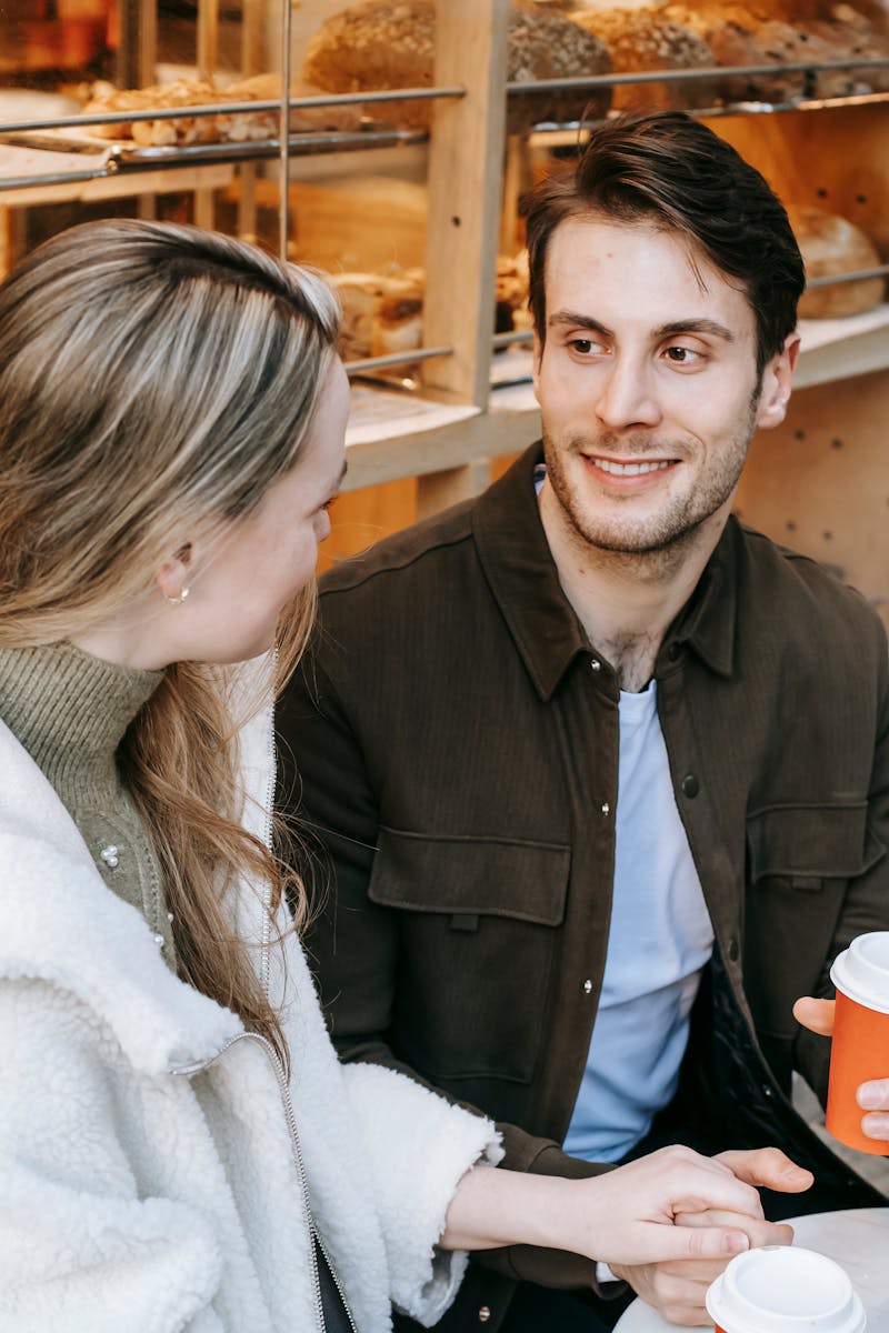 Positive young boyfriend and girlfriend sitting at table in bakery and holding hands while talking and enjoying takeaway coffee