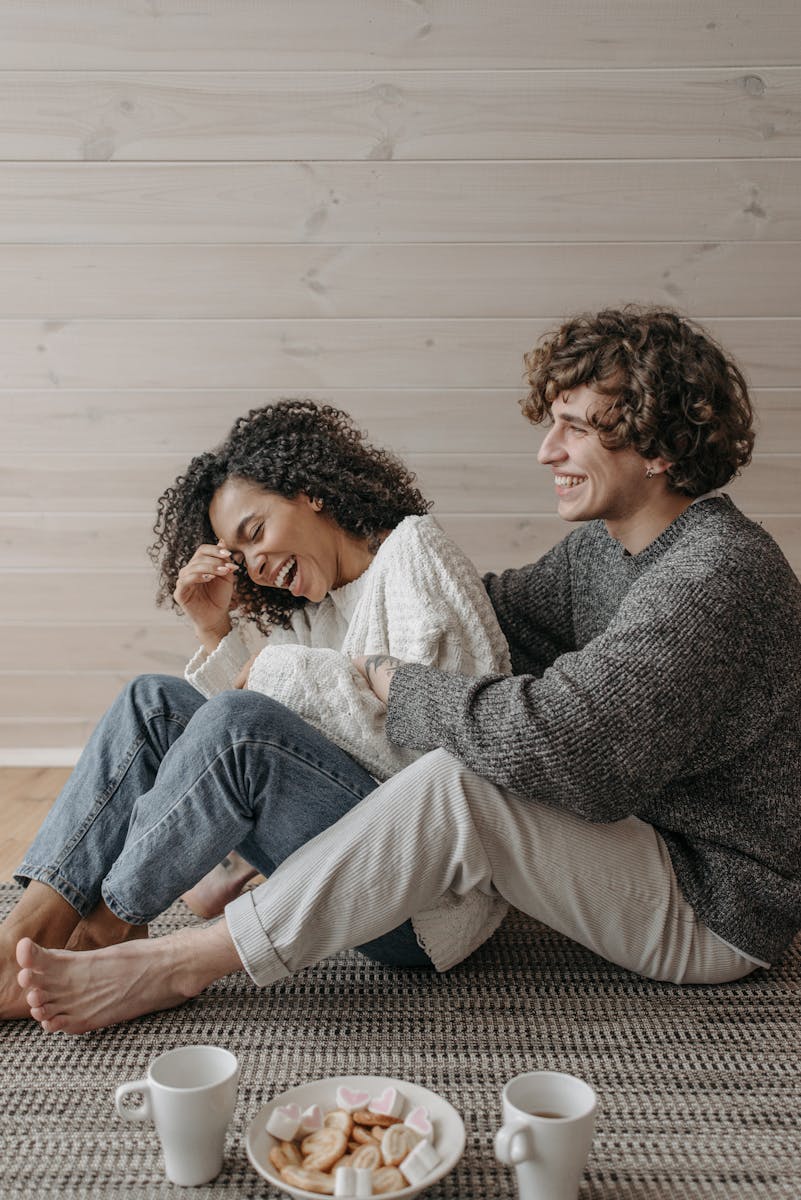A joyful couple sitting on the floor, enjoying a relaxed moment indoors, wearing knitted sweaters, with snacks and coffee cups.