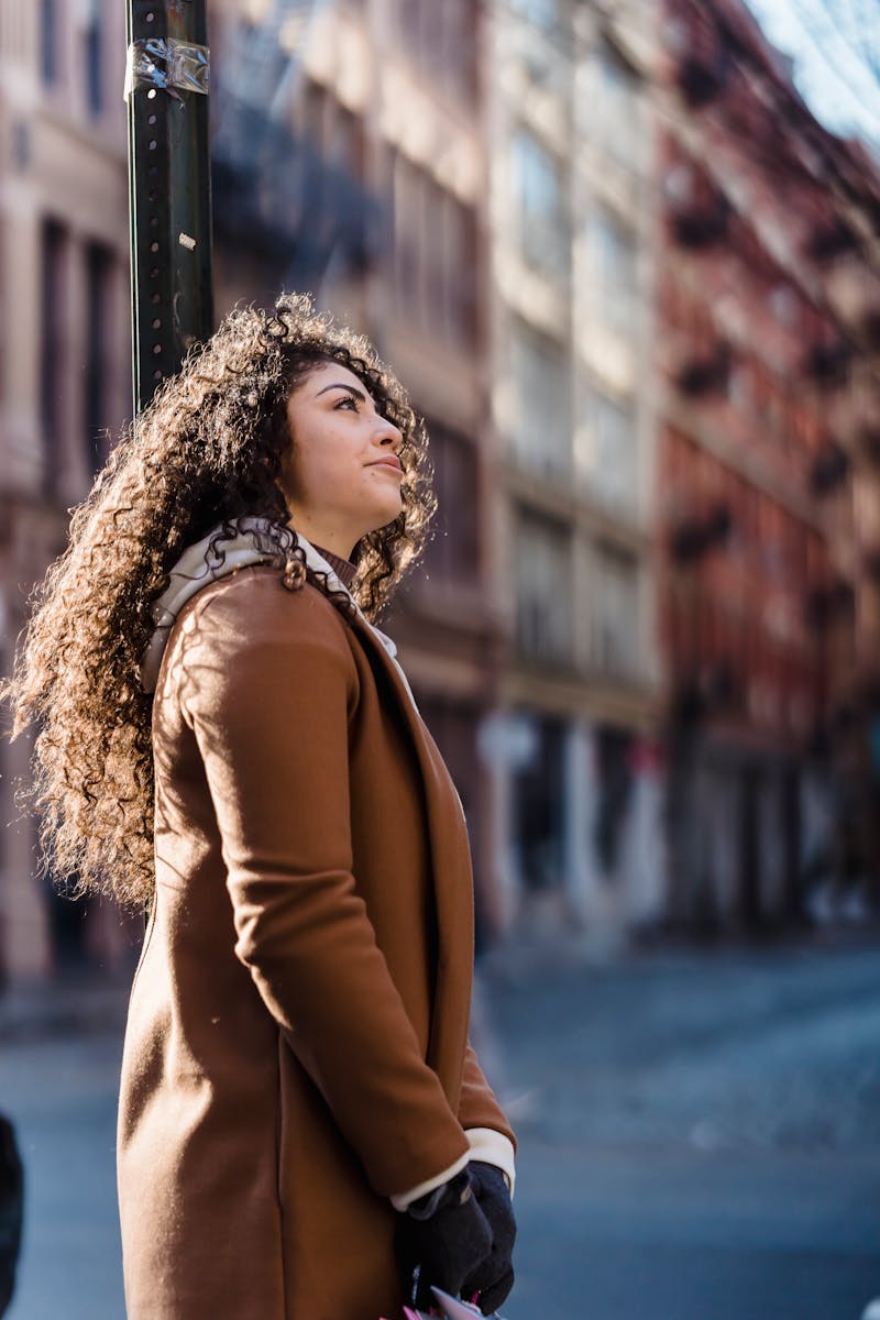Side view of young woman wearing warm coat while standing in street near road and building in town in sunny day