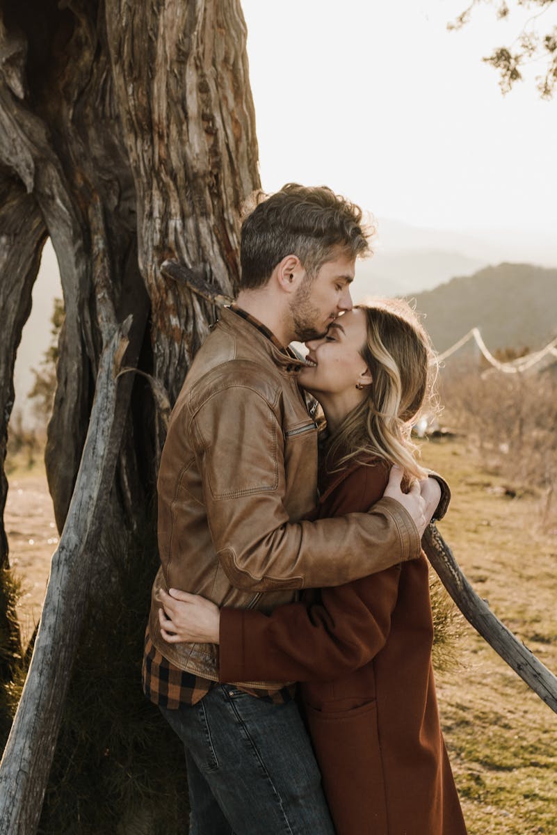 A romantic couple embraces outdoors by a tree trunk, bathed in warm sunlight, showcasing love and connection.