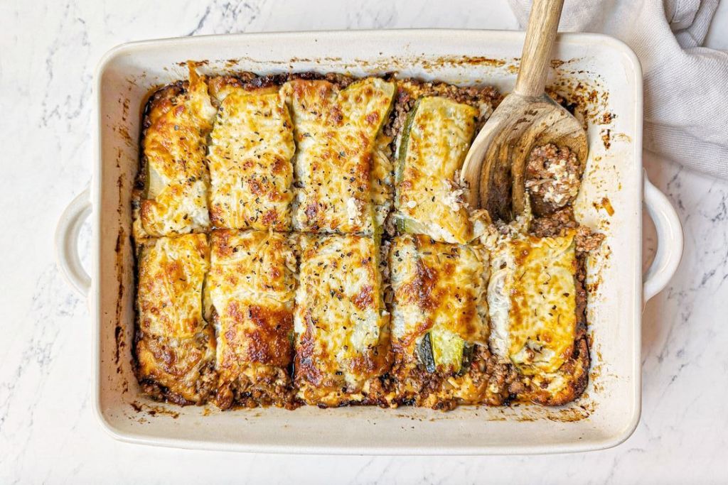 A baked zucchini lasagna dish featuring layers of zucchini, ground beef, marinara sauce, ricotta, and melted mozzarella cheese, presented in a baking dish with a wooden spoon on the side.