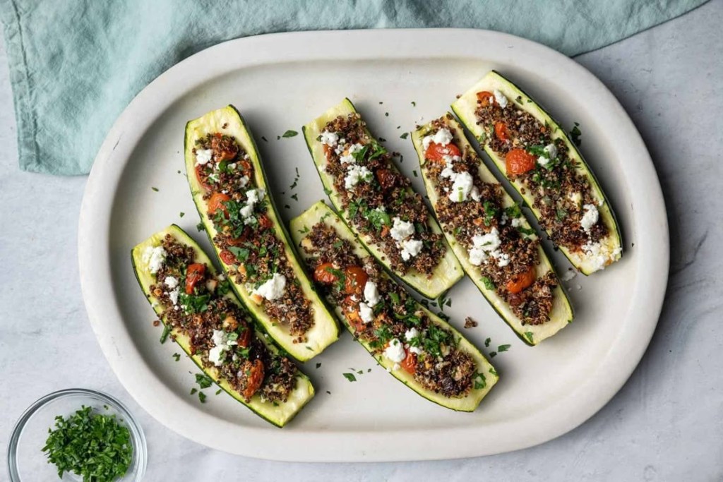 A platter of stuffed zucchini boats filled with quinoa, cherry tomatoes, and crumbled feta, garnished with fresh herbs.