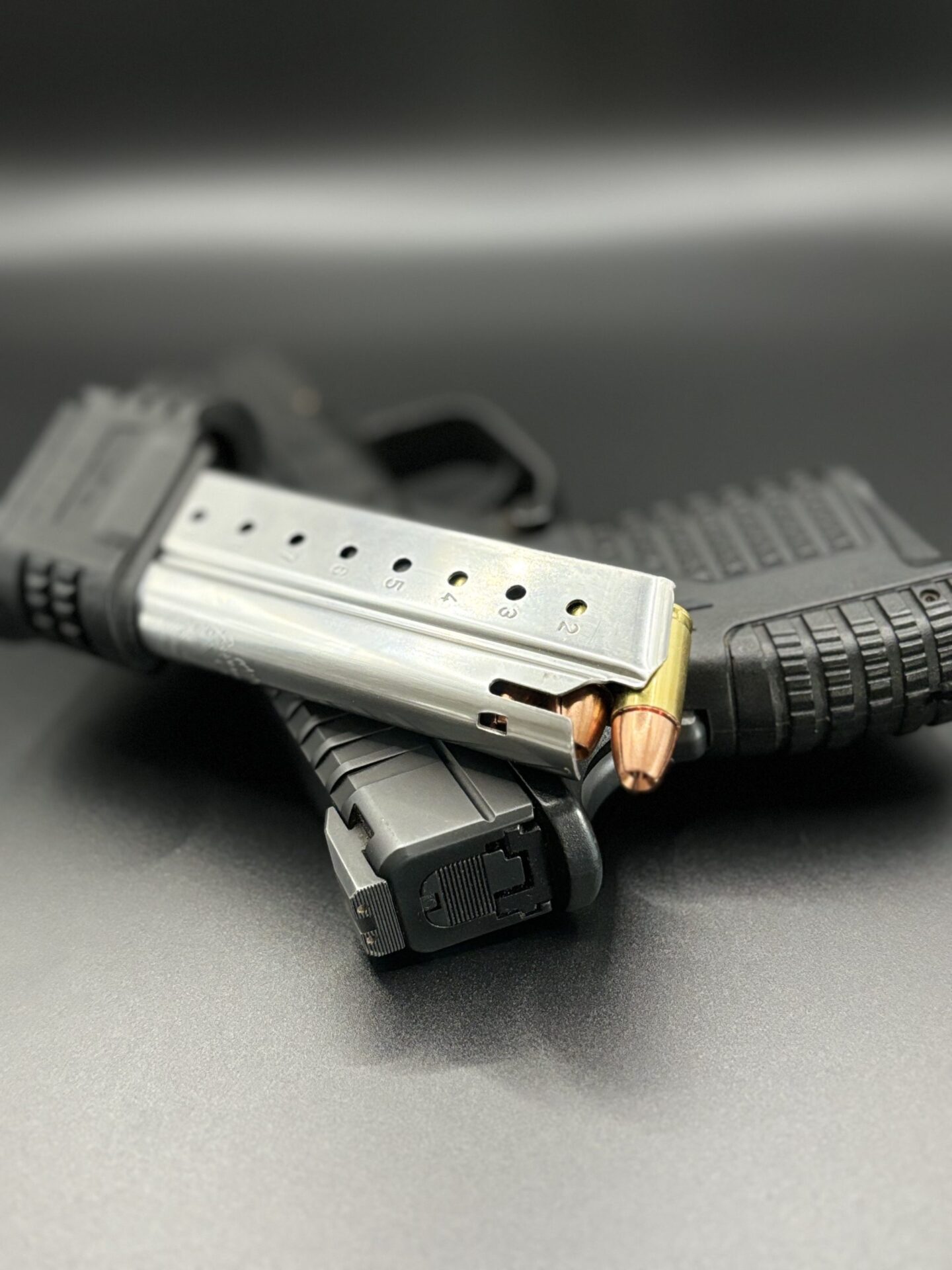 ms Ammunition – High-Quality Ammunition for Rifles and Shotguns" A pistol is positioned on a smooth surface alongside two magazines, one of which is loaded with visible high-quality ammo. The emphasis is on the magazine and rifle cartridges, providing a clear view of the ammunition. Meanwhile, shotgun shells are subtly highlighted as part of the firearm's components in the softly blurred background. This scene effectively illustrates an assortment of bulk ammunition options for various firearms. Bulk Ammo Available for Purchase"