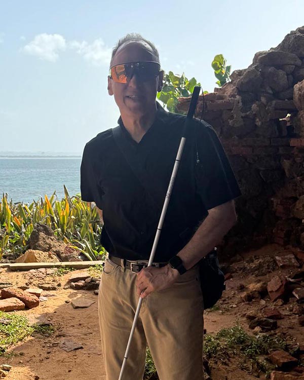 A photo of a Blind Latin American man wearing sunglasses and holding a white cane. He is standing in front of a large body of water.
