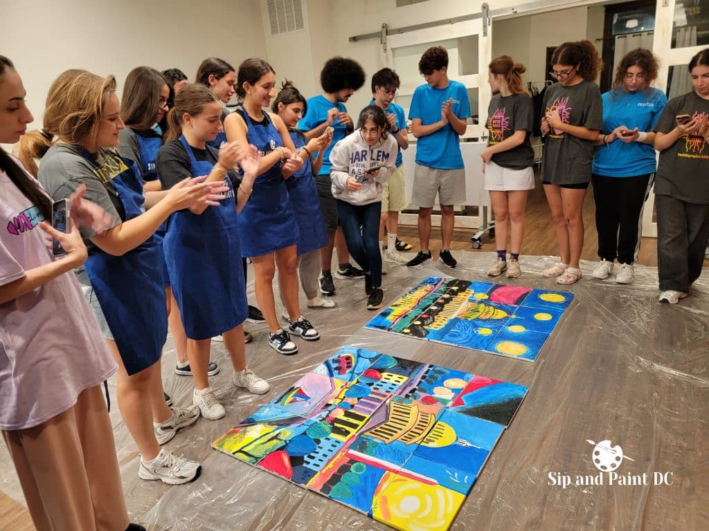 A group of people in a room at Sip and Paint DC, viewing two large, colorful paintings on the floor. Some are clapping, and others observe quietly.