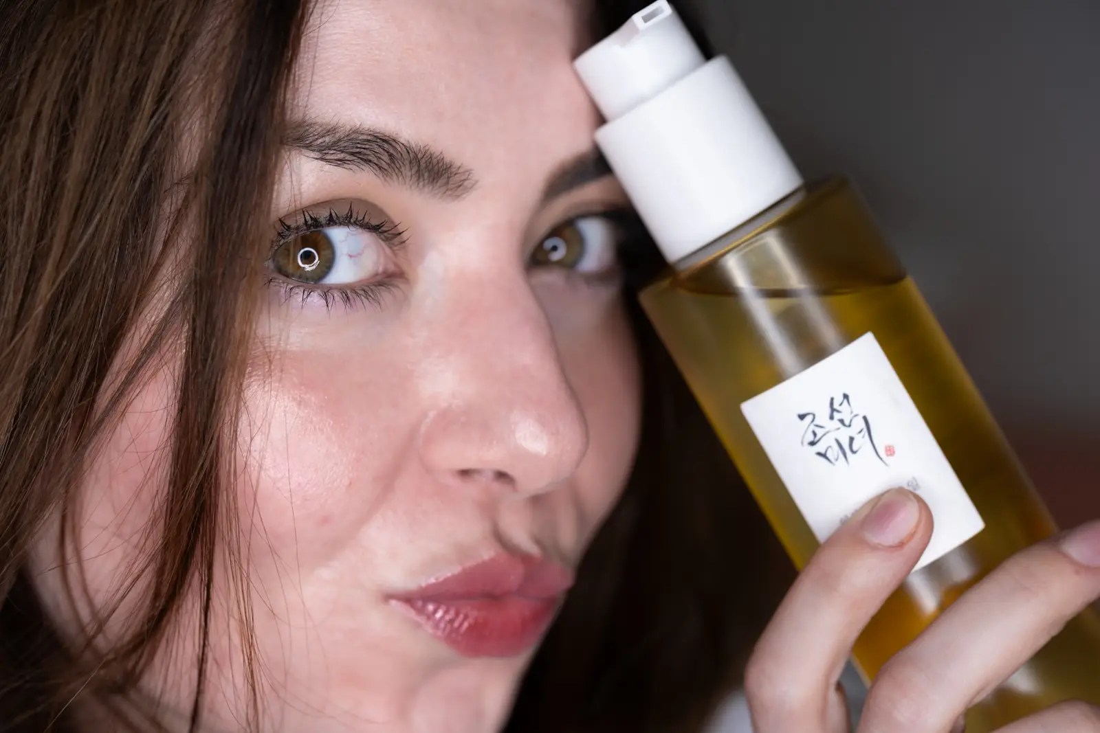 Close-up of a young woman with fair skin and green-hazel eyes holding a bottle of Korean skincare product with Korean writing on the label. She has natural makeup, full pink lips, and chestnut brown hair