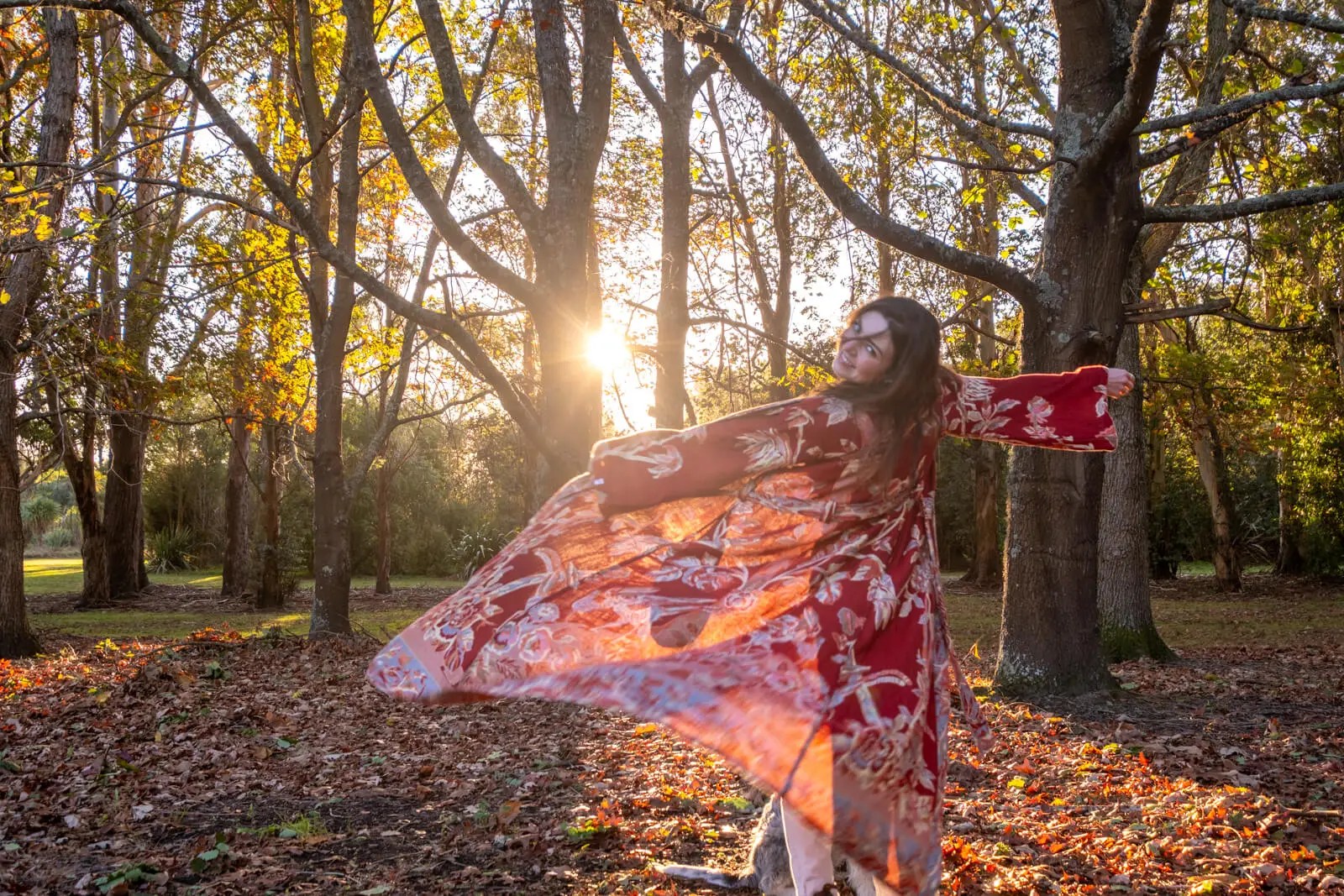 Woman twirling in a flowing red floral kaftan in an autumn forest at golden hour, with a soft smile and sunlight filtering through the trees