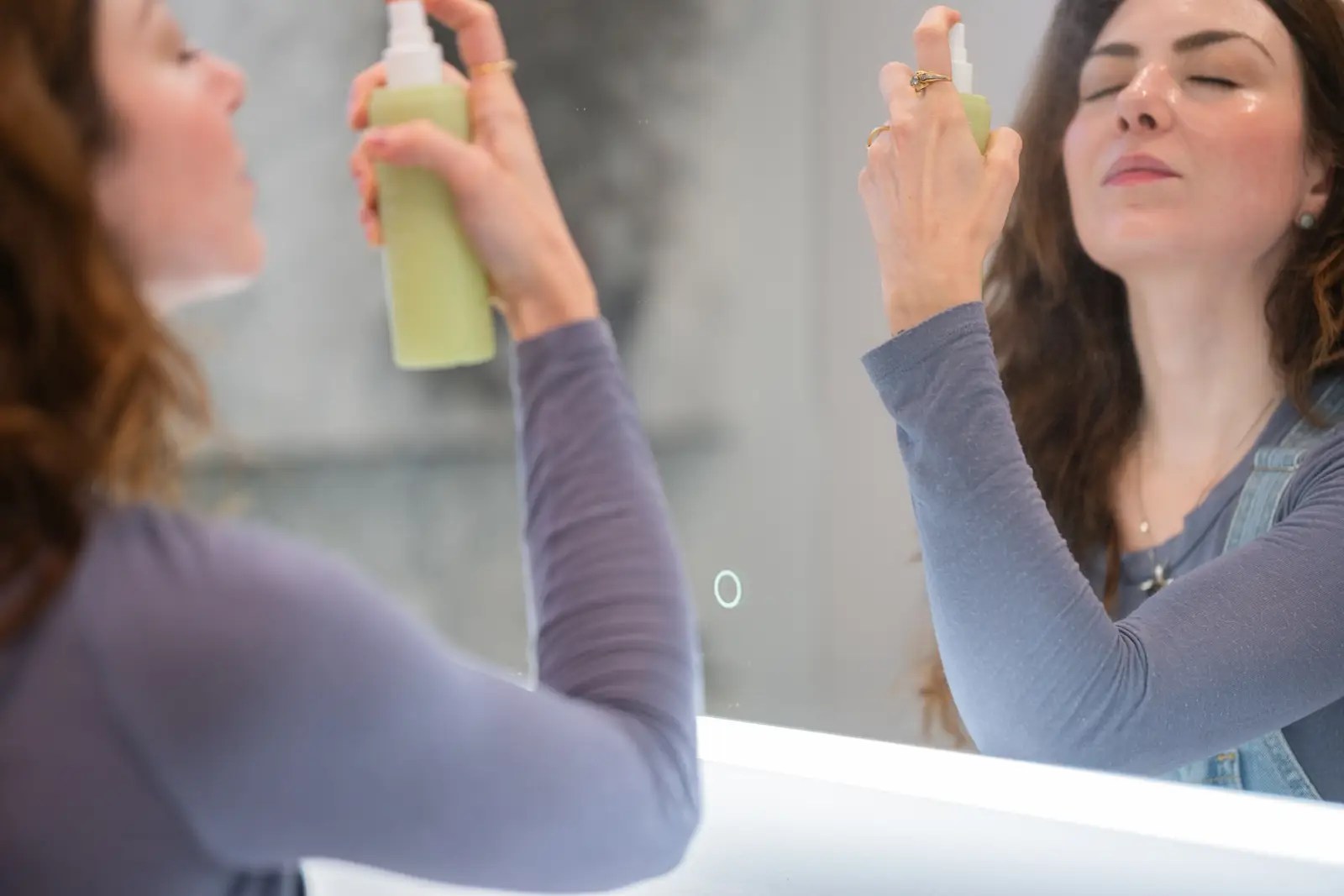 Woman applying facial mist in front of a bathroom mirror as part of her skincare routine, holding a green spray bottle with eyes closed and wearing a long-sleeve shirt and necklace