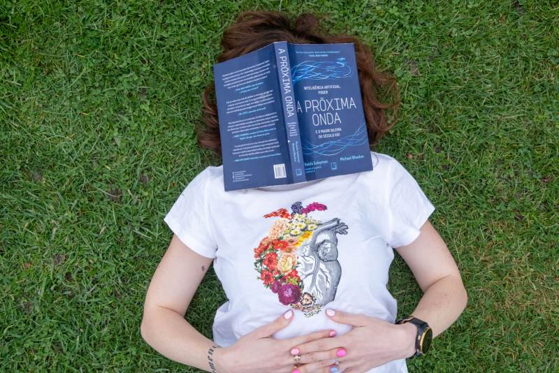 Person lying on the grass with the book "The Coming Wave" by Mustafa Suleyman and Michael Bhaskar covering their face. They are wearing a white t-shirt with an artistic heart and flower design. The scene symbolizes reflection on the future of technology and artificial intelligence.