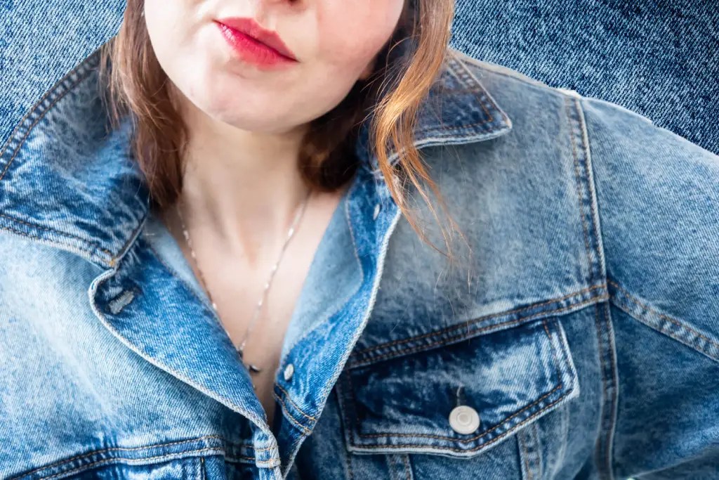 Close-up of a woman in a vintage blue denim jacket with the collar popped, wearing a red lipstick and a delicate silver necklace, against a denim-textured background.
