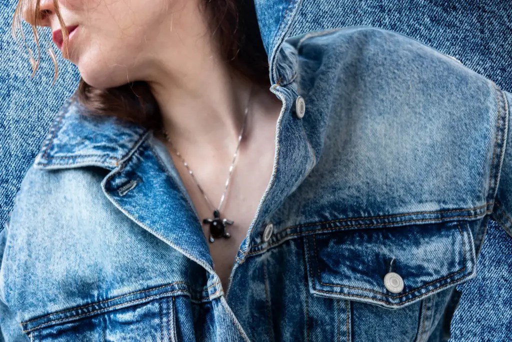 Close-up of a woman turning her head while wearing a faded blue denim jacket and a black turtle pendant necklace, with a dark denim backdrop.
