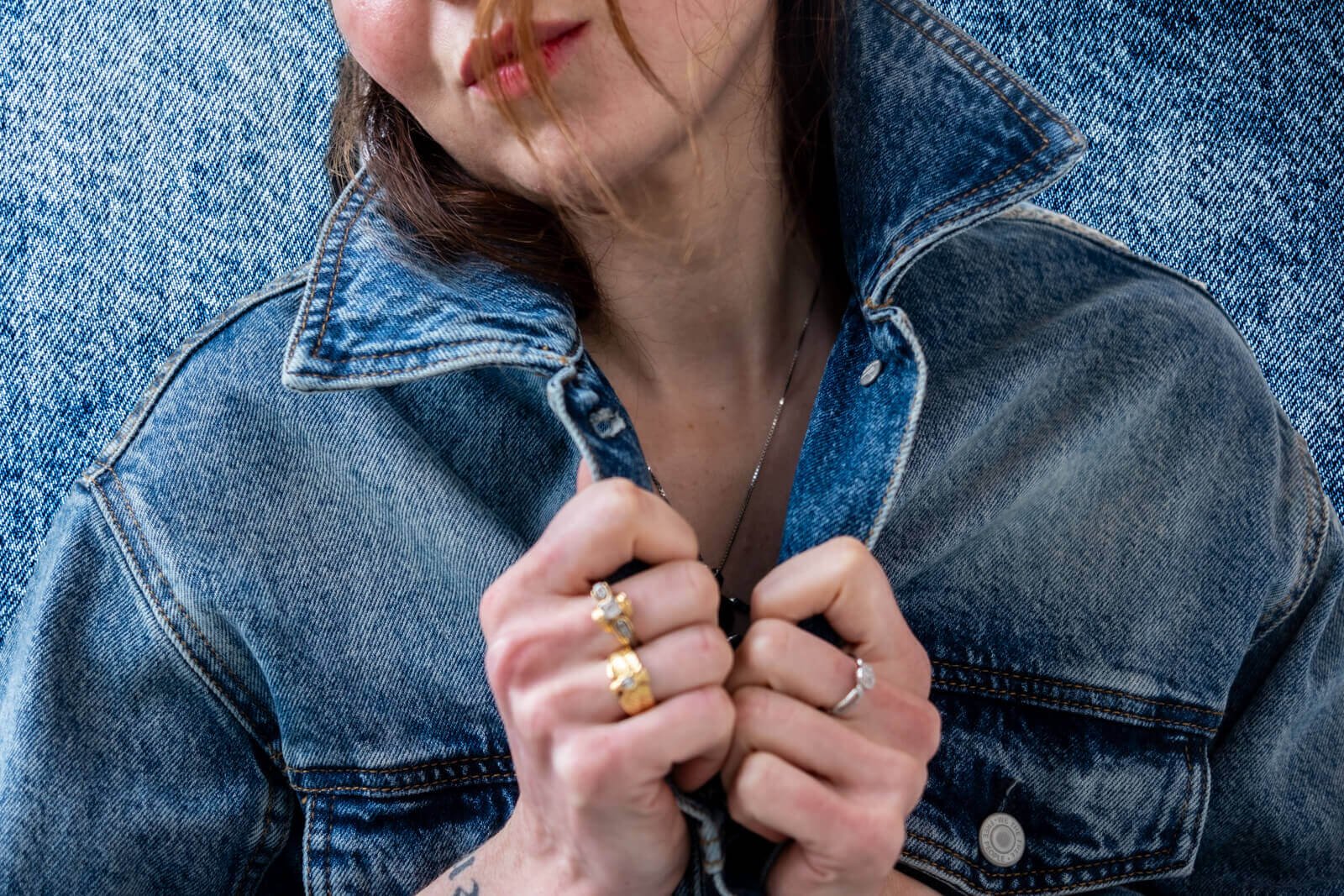 Close-up of a woman wearing a denim jacket, holding the collar with both hands. She is accessorized with multiple rings and a necklace. The background is also denim, creating a monochromatic, textured look. Her face is partially visible with loose hair strands and a subtle expression.